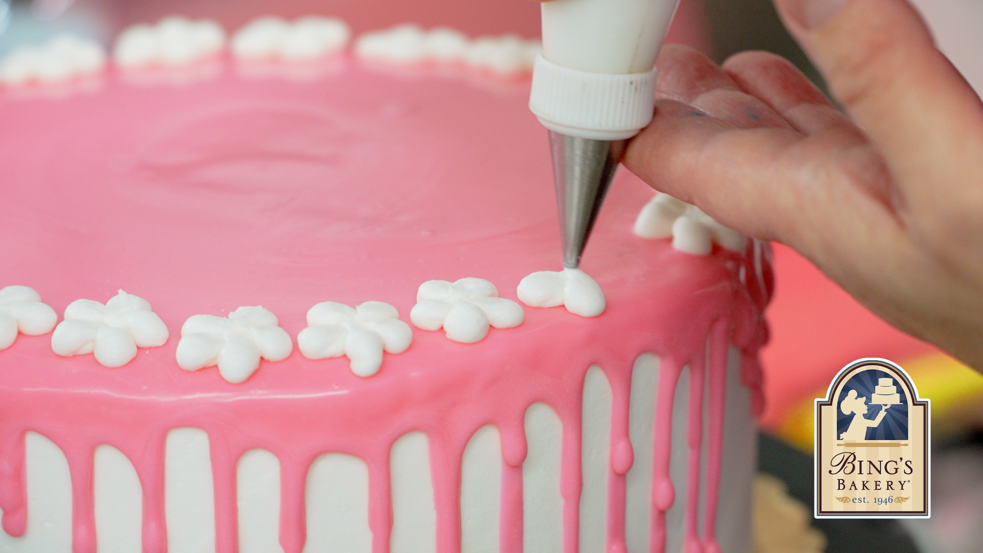 Baker piping white icing flowers onto a pink drip cake at Bings Bakery